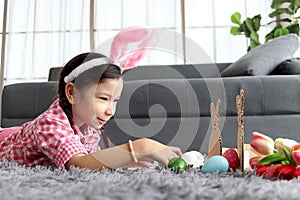 Happy adorable pink little Easter bunny girl kid with rabbit ears headband laying down with colorful painted Easter eggs in living