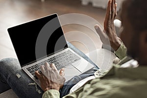 Happy adult black man using computer with blank screen, waving hand at webcam, have meeting, sit on sofa