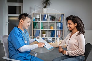 Happy adult chinese man doctor showing contract to young african american female patient in clinic