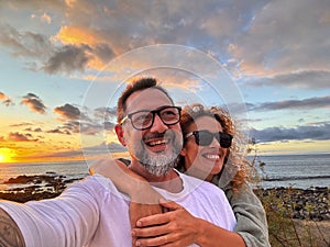 Happy adult couple in love taking selfie picture with romantic wonderful sunset on the ocean in background. Young mature people