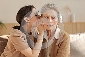 Happy adult daughter whispering secret to her middle mother at home, gossiping, sharing secret