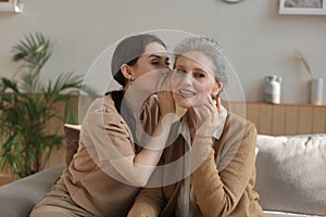 Happy adult daughter whispering secret to her middle mother at home, gossiping, sharing secret