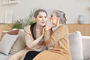 Happy adult daughter whispering secret to her middle mother at home, gossiping, sharing secret