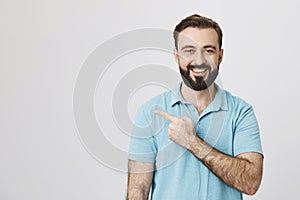 Happy adult european man smiling cheerfully while pointing left with index finger, over gray background. Guy in theme