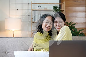 Happy adult granddaughter and senior grandmother having fun enjoying talk sit on sofa in modern living room, smiling old