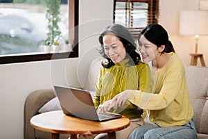 Happy adult granddaughter and senior grandmother having fun enjoying talk sit on sofa in modern living room, using a laptop,