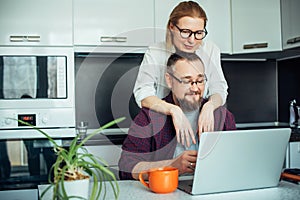 Happy adult loving family couple looking at laptop screen, sitting at table in kitchen. Bearded man showing funny video on