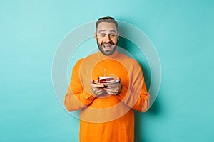 Happy adult man celebrating birthday, holding bday cake with candle and smiling, standing against turquoise background