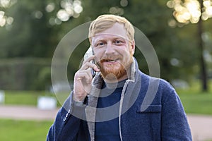 Happy adult red haired man blue eyes, beard, mustache smiling, talking smartphone, mobile. stylish young ginger bearded guy using