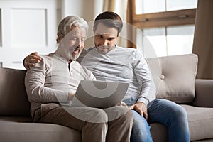 Happy adult son and senior dad use laptop at home