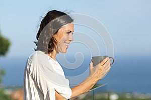 Happy adult woman with coffee looking away in a balcony