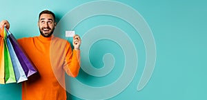 Happy aduly man showing credit card and shopping bags, standing against turquoise background