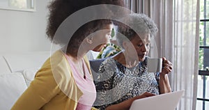 Happy african american adult daughter and senior mother using laptop at home, slow motion