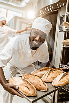 happy african american baker taking bread loaves from oven