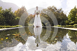 Happy african american bride holding bouquet, reflected in pond, standing in sunny garden