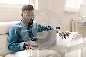 Happy african american businessman sitting in office on couch, working on laptop computer and smiling, copy space