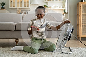 Happy African American child boy relaxing with smartphone in front of electric fan at home