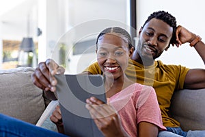 Happy african american couple smiling and embracing and using tablet on couch at home