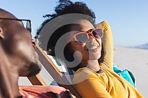 Happy african american couple in sunglasses sitting in deckchairs smiling on sunny beach