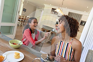 Happy african american daughter feeding mother sitting at table during breakfast and smiling