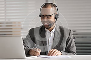 Happy African American employee in headset making notes, using laptop