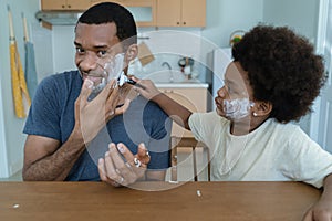 Happy African American Father and little son having fun while shaving and looking at mirror with kid holding razor helping to