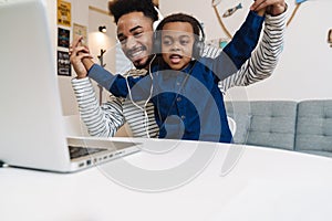 Happy african american father and son in headphones using laptop at home