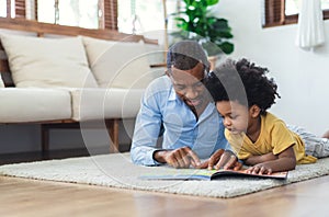 Happy African American father and son are reading a book and smiling while lying on floor spending time together at home. Children