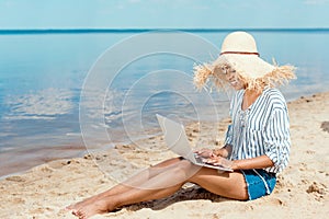 happy african american female freelancer in straw hat sitting on sandy beach