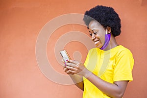 Happy African-American female with a medical mask using a modern smartphone checking social media