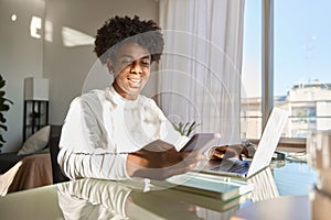 Happy African American gen z teen student using mobile phone and laptop at home.