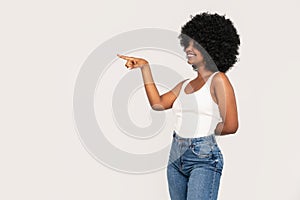 Happy african-american girl with afro hairstyle and big smile pointing on the empty studio background. Young woman in white top