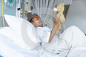 Happy african american girl patient lying on bed playing with teddy bear in patient room at hospital