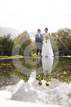 Happy african american groom and bride holding bouquet reflected in pond in sunny garden, copy space