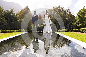 Happy african american groom and bride holding bouquet reflected in pond in sunny garden, copy space