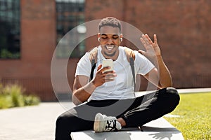 Happy african american guy making video call on cellphone, waving hand to webcamera while walking in the city