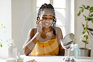 Happy african american lady looking at mirror and applying under eye cream, sitting wrapped in towel at home