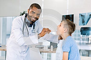 Happy African American male doctor watching patient boy with sore throat in clinic. The doctor examining little boy`s
