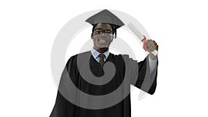 Happy african american male student in graduation robe walking with diploma and talking to camera on white background.