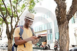 Happy african american man with bag and hat walking with mobile phone outdoors