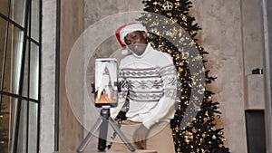 Happy african american man in christmas hat dancing energetically in living room in front of smartphone