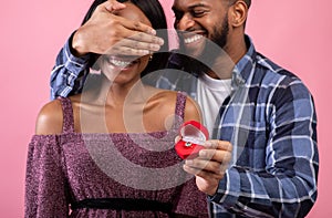 Happy African American man covering his fiancee`s eyes, holding box with engagement ring on pink background