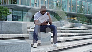 Happy African American man euphoric winner with smartphone on street in city. Person guy looking at cell phone reading