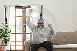 Happy african american man having fun at home, screaming with remote controller in his hand, watching football game on