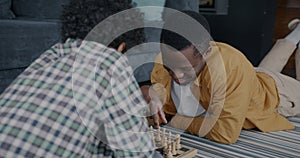 Happy African American man playing chess with little son concentrated on game smiling lying on floor at home