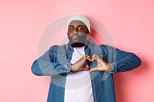 Happy african-american man showing heart sign, I love you gesture, pucker lips for kiss while standing over pink