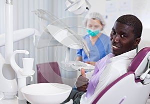 Happy african american man sitting in dental chair after teeth cure giving thumb up