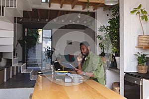 Happy african american man sitting at table in kitchen using laptop and talking on smartphone