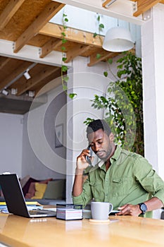 Happy african american man standing at table in kitchen, using laptop and talking on smartphone