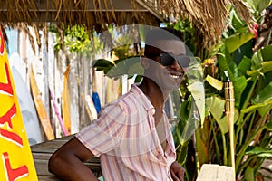 Happy african american man in sunglasses leaning on counter at surf hire beach shack
